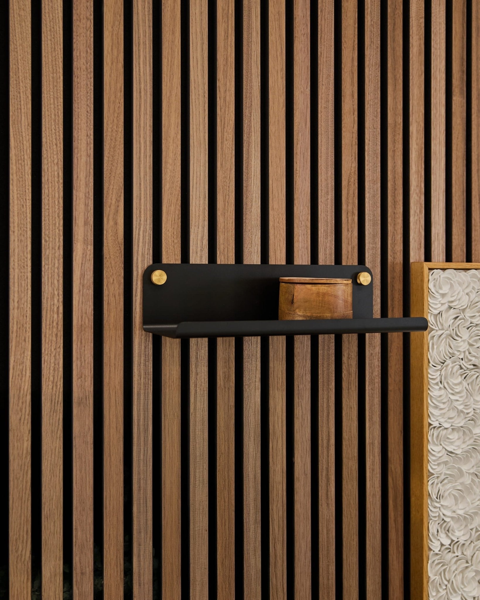 Una piccola scatola di legno su una mensola WoodUpp Norr Wide (nera, 35,9 cm) fissata a doghe acustiche verticali in legno Akupanel, con un pannello bianco ruvido in evidenza.